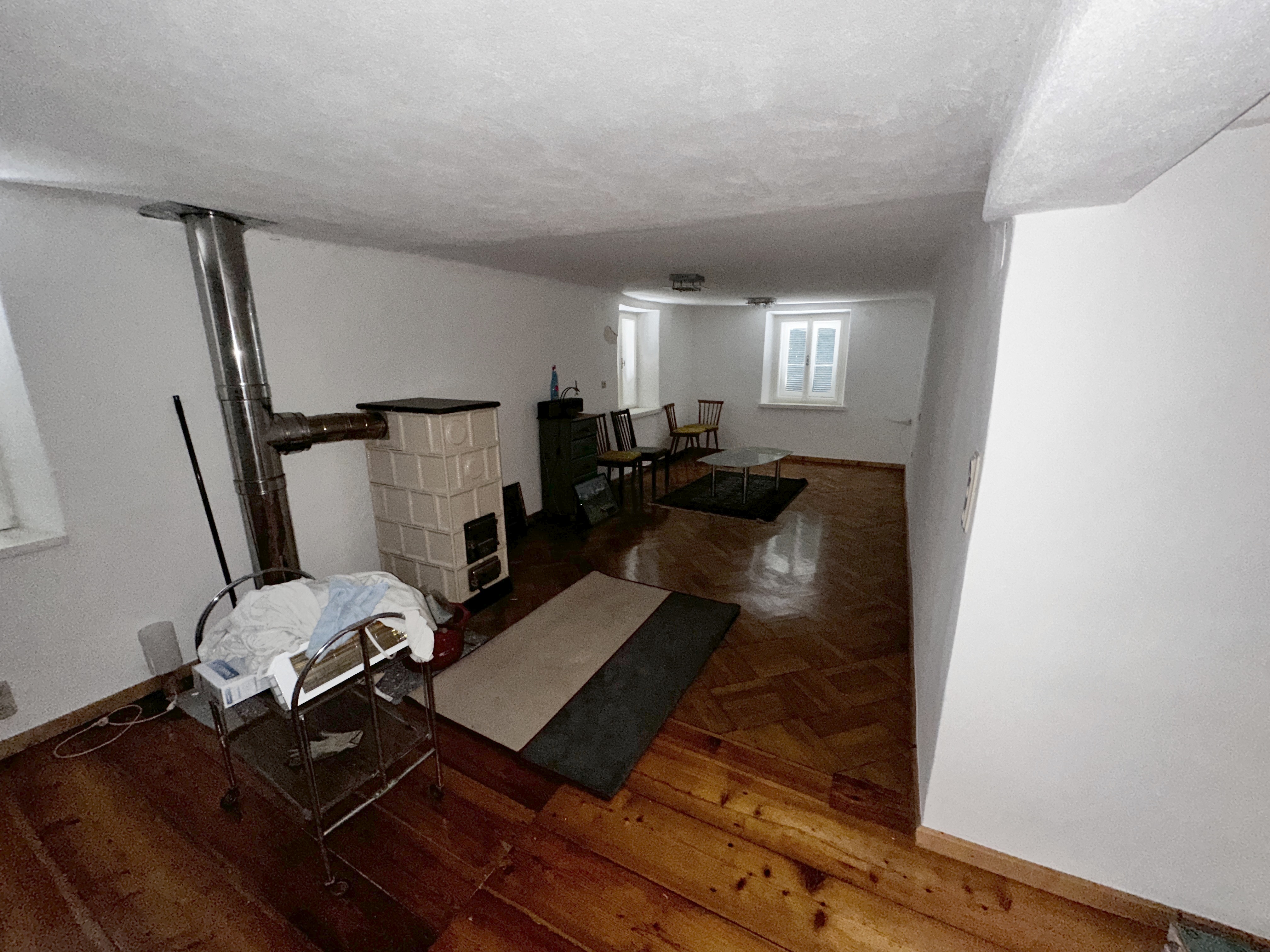 Aufgehellter Wohnraum mit Kachelofen, Edelstahlrohr, Holzfußboden und vorhandenem Mobiliar.